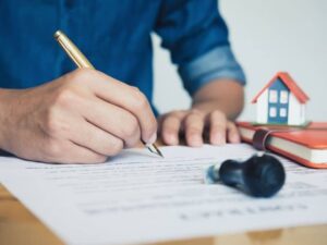 Person signing property documents with house model on the desk representing elevation certificate and home ownership verification for real estate records.